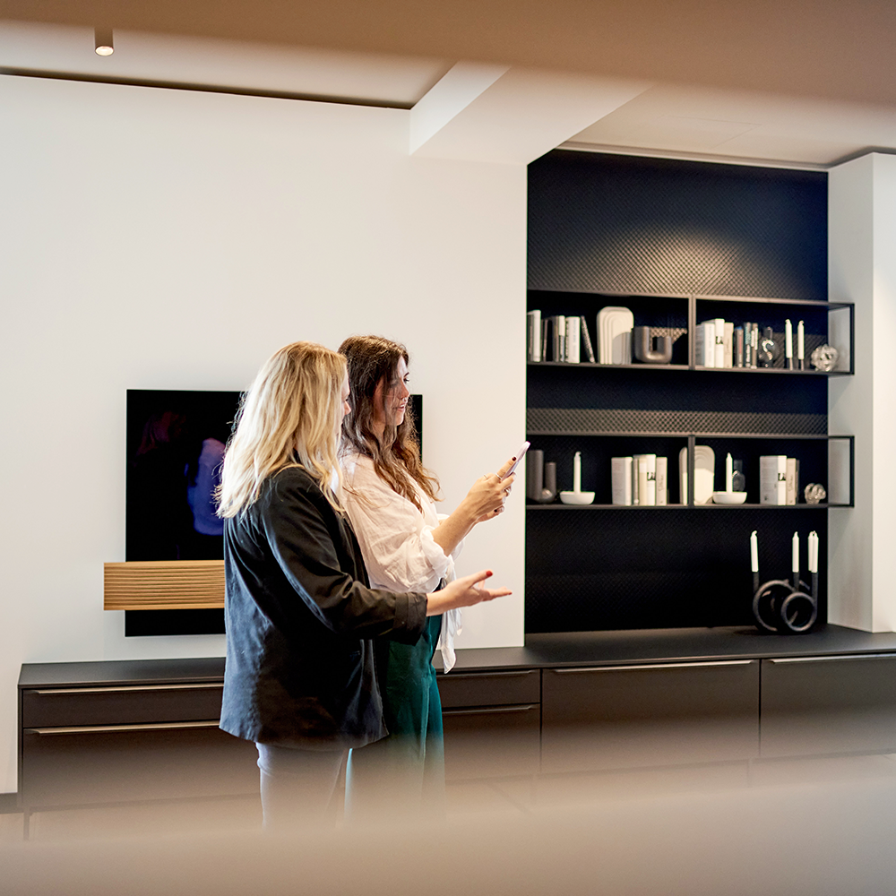 Auf dem Bild sieht man zwei redende Frauen vor einer TV-Wand mit zwei Regalen und vier Unterschränken im Wohnzimmer, welche gemeinsam auf ein Handy schauen.